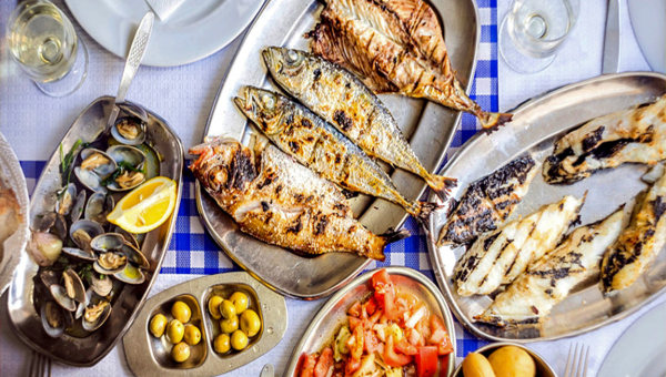 Assorted grilled fish, shellfish, olives, and salad on metal trays on a table.