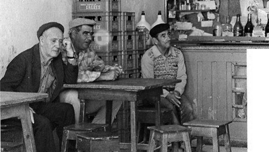 Three men in a vintage cafe, sitting at tables, with bottles and shelves in the background.