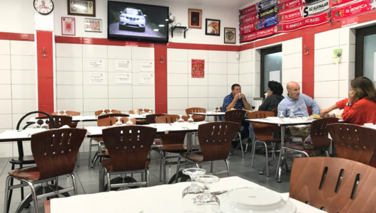 Casual restaurant interior with diners, white walls, red accents, and a wall-mounted TV.