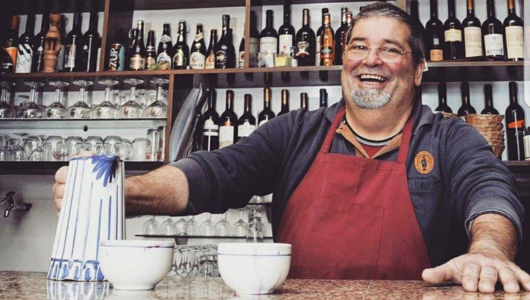 Smiling man in a red apron behind a bar with bottles and glasses, holding a jug.