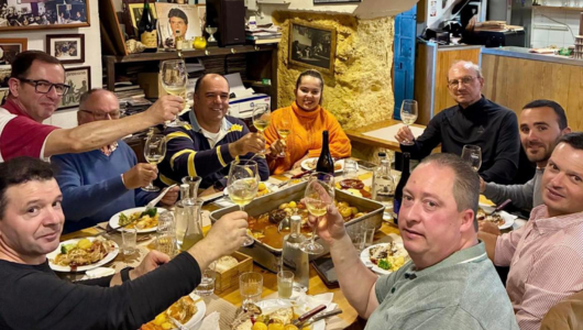 Eight people seated at a dinner table raising wine glasses in a toast.