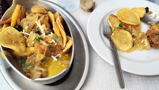 Dishes with fried potatoes, onions, and herbs on a table with a fork and bread.