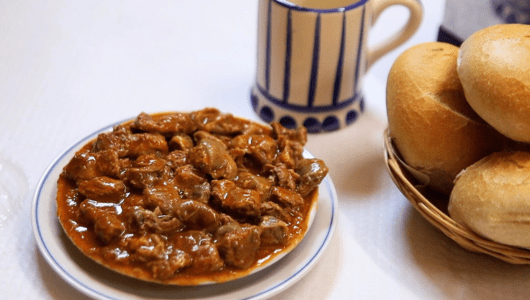 Plate of meat stew beside a basket of bread rolls and a striped mug.
