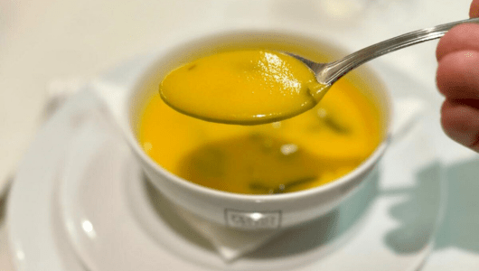 Spoon with yellow soup above a white bowl filled with soup on a plate.