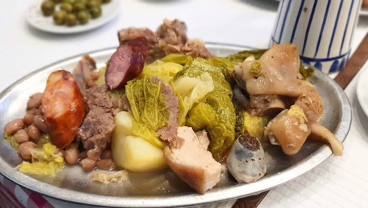 Plate of mixed meats, beans, cabbage, and potatoes on a table with a cup and olives in the background.