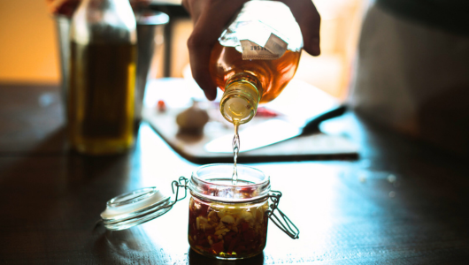 Hand pouring oil into a glass jar with chopped ingredients on a wooden table.