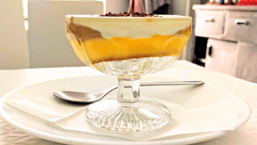 Glass dessert cup with layered custard and whipped cream on a plate with a spoon.
