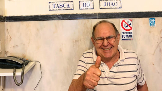 Smiling man giving thumbs up in front of a sign reading 'TASCA DO JOAO' with a 'No Smoking' sign nearby.