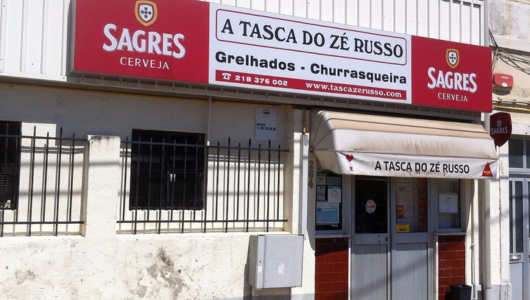 Restaurant entrance with signs 'A Tasca do Zé Russo' and 'Sagres Cerveja' on a sunny day.
