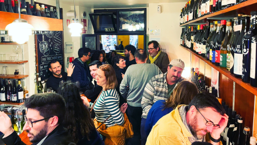 Crowded wine bar with people socializing and bottles on shelves.