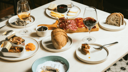 Charcuterie board with wine, bread, cheese, oysters, and tinned fish on a table.