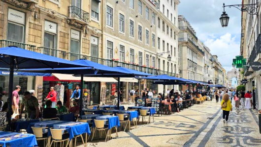 Outdoor cafe with blue umbrellas on a bustling street with tiled pavement and historic buildings.