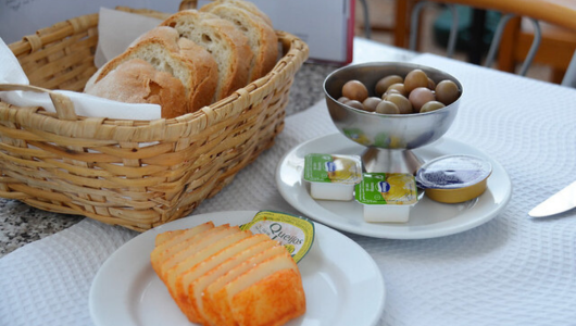 Bread basket, sliced cheese, olives, and butter portions on a table.