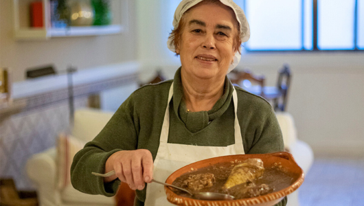 Person holding a dish of food with a spoon, wearing an apron and cap in a cozy indoor setting.