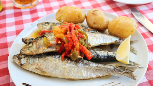 00.A brief guide to ordering fish in Portugal_feat photo Grilled sardines with roasted peppers and potatoes on a white plate with a lemon wedge, on a red checkered tablecloth.