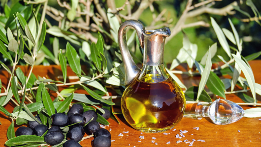 Glass jug of olive oil with black olives and olive branches on a wooden table.