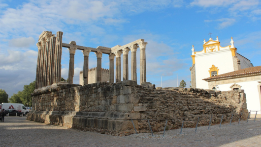 Roman temple ruins with columns beside a white church under a cloudy sky.