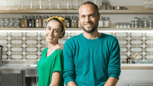 Two smiling people in casual clothes standing in a modern kitchen with shelves and jars.