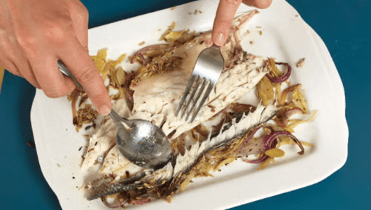 Hands removing fish bones with fork and spoon on a plate.