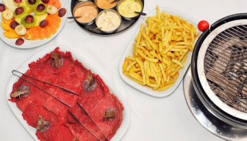 Assorted fruits, sauces, fries, and raw beef slices on a table with a grill.