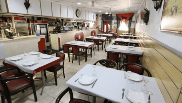 Empty restaurant with red chairs, white tablecloths, and a visible kitchen counter.