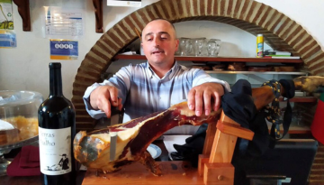 Man slicing cured ham on a wooden stand next to a wine bottle indoors.