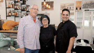 Three people standing in a restaurant, two in chef attire, smiling at the camera.