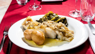 Plate with fish, potatoes, and greens on a red tablecloth.