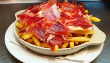 Fries topped with thin slices of cured meat in a bowl on a plate.