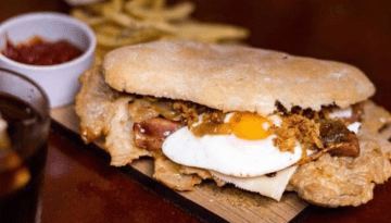 Sandwich with fried egg, meat, and toppings on a ciabatta, with fries and sauce on the side.