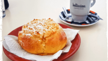 Pastry on red plate next to a 'bellissimo' cup on saucer with spoons.