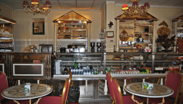 Interior of a small cafe with ornate decor, tables, chairs, and a counter displaying drinks and snacks.