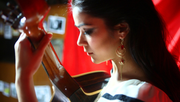 00.How to experience Fado in Lisbon_feat photo Woman holding a guitar against a red backdrop, wearing earrings and a striped top.