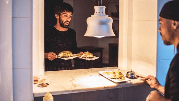 00.Óbidos travel guide for food lovers_feat photo Two men exchanging plates of food through a kitchen serving hatch.