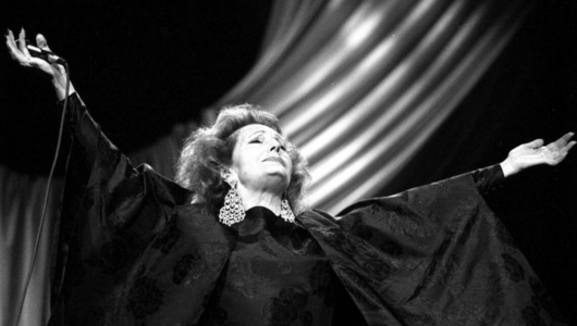 Person singing on stage with arms raised, wearing large earrings and a flowing outfit.