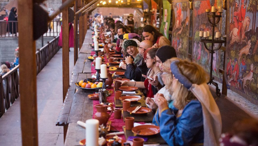 People in medieval attire dining at a long wooden table with candles and fruit.