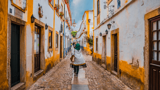 Person walking down a narrow, colorful street with cobblestones and yellow-trimmed buildings.