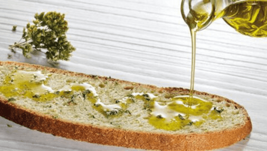 Olive oil being poured onto a slice of bread with herbs on a white wooden surface.