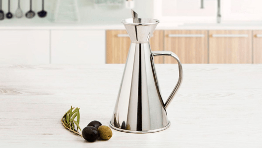 Metal oil dispenser with handle beside black and green olives on a kitchen counter.