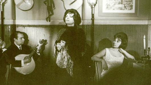 Singer performing with a guitarist; a woman is seated looking on in a vintage setting.