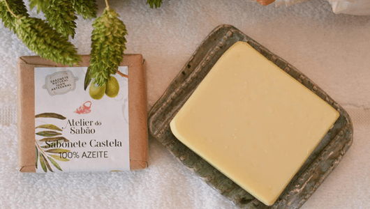 Soap bar on green dish next to wrapped soap labeled 'Atelier do Sabao' on a towel.
