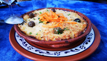 Baked dish with grated cheese, olives, and carrot strips in a ceramic bowl on a decorative plate.