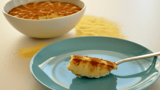 A spoon with pudding on a plate, bowl with cinnamon pudding in background.