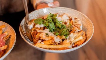 Bowl of fries topped with cheese and green herbs, held by a person.