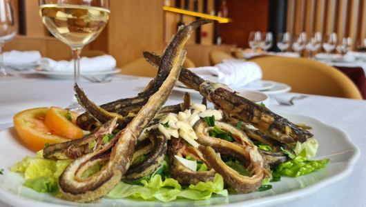 Fried eels on a bed of greens and tomatoes, with a glass of white wine in a restaurant setting.