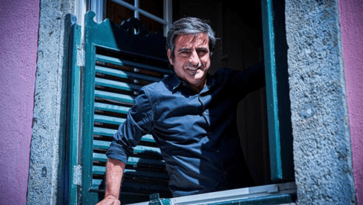 Person in blue shirt smiling while leaning out of a window with a green shutter.