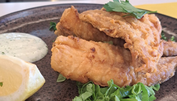 Fried fish on greens with lemon wedge and herb sauce on a dark plate.