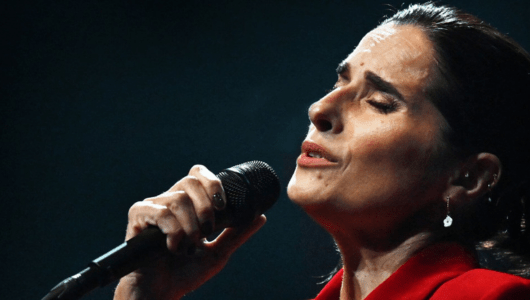 Person in red shirt singing passionately into a microphone against dark background.