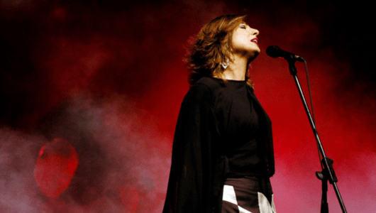 Woman singing passionately on stage with a microphone, red smoke in background.