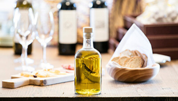 Bottle of olive oil on a table with bread, wine bottles, and glasses in the background.